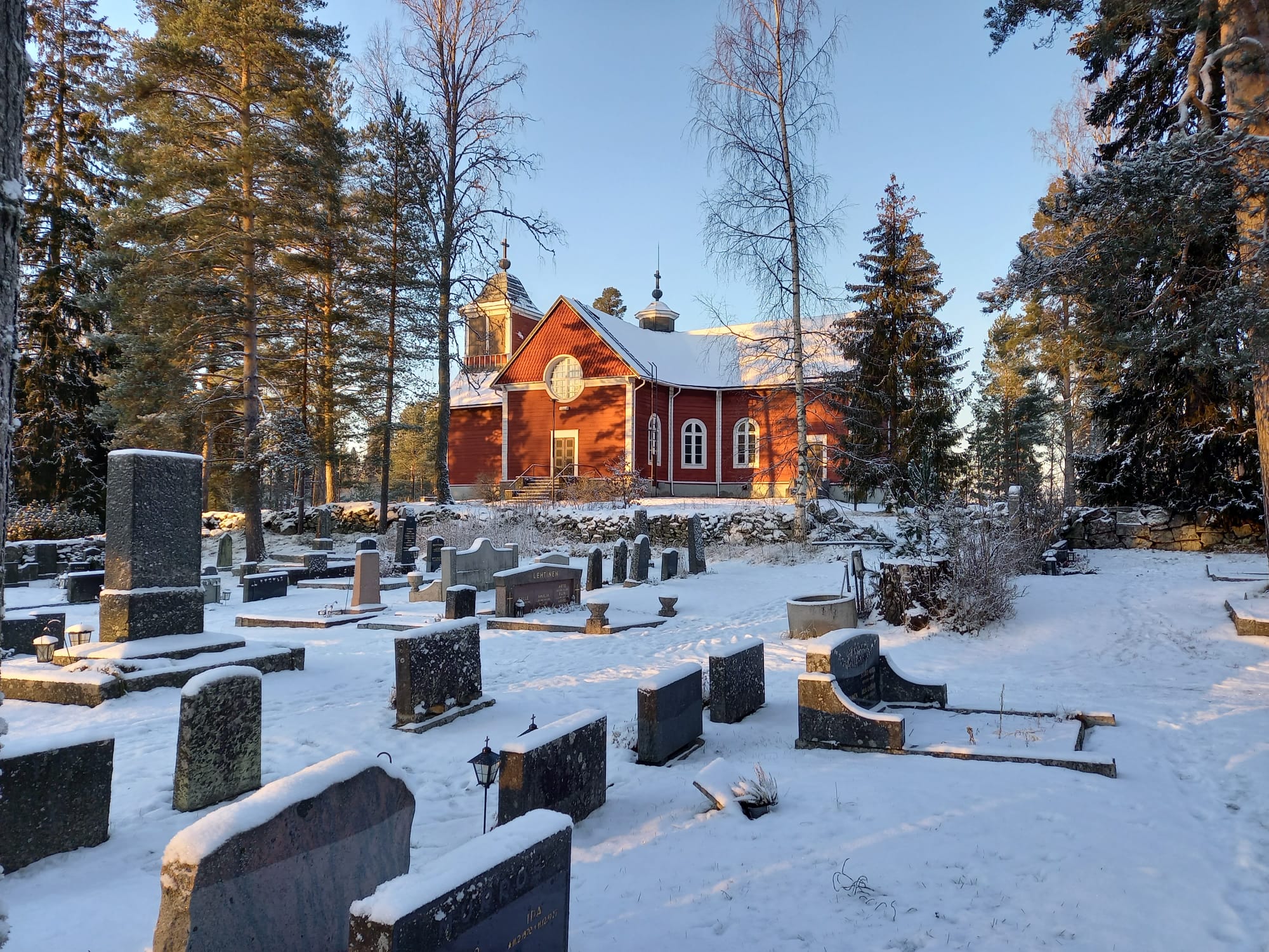 Pukkilan punainen kirkko hohtaa auringon valossa, etualalla hautakiviä ja hautausmaan puustoa.